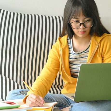 Asian female student wearing glasses is researching with a laptop and taking notes.